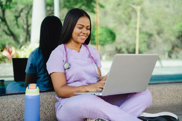 a woman in purple nurse scrubs sits on the ground and works on a laptop.