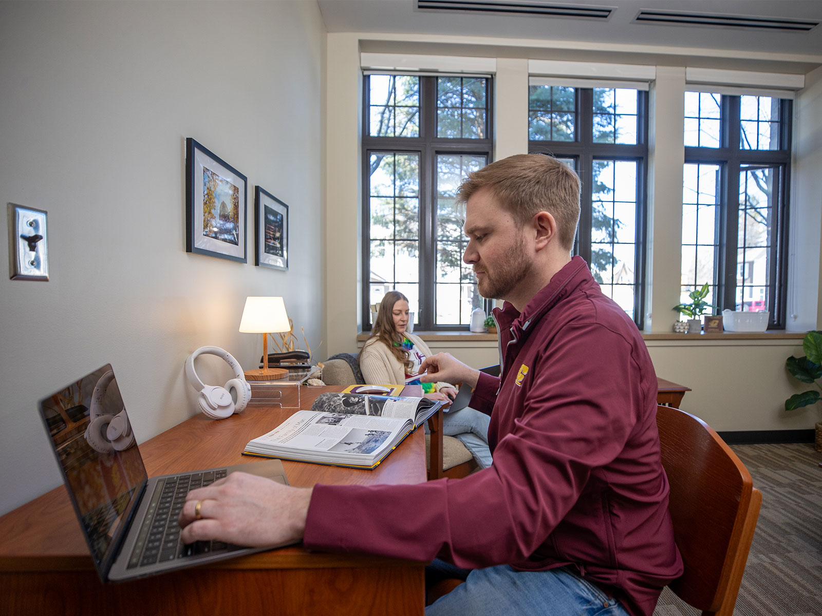 A man wearing a CMJ jacket sits at a desk with a laptop and a book nearby is a woman by a window with a laptop