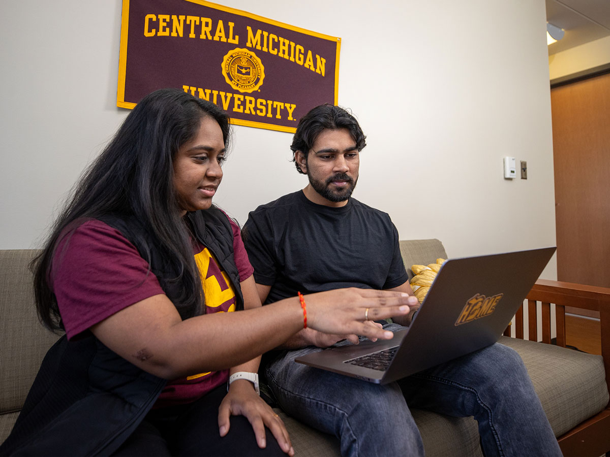 Two students sitting on a couch looking at a laptop with a Central Michigan University banner behind them