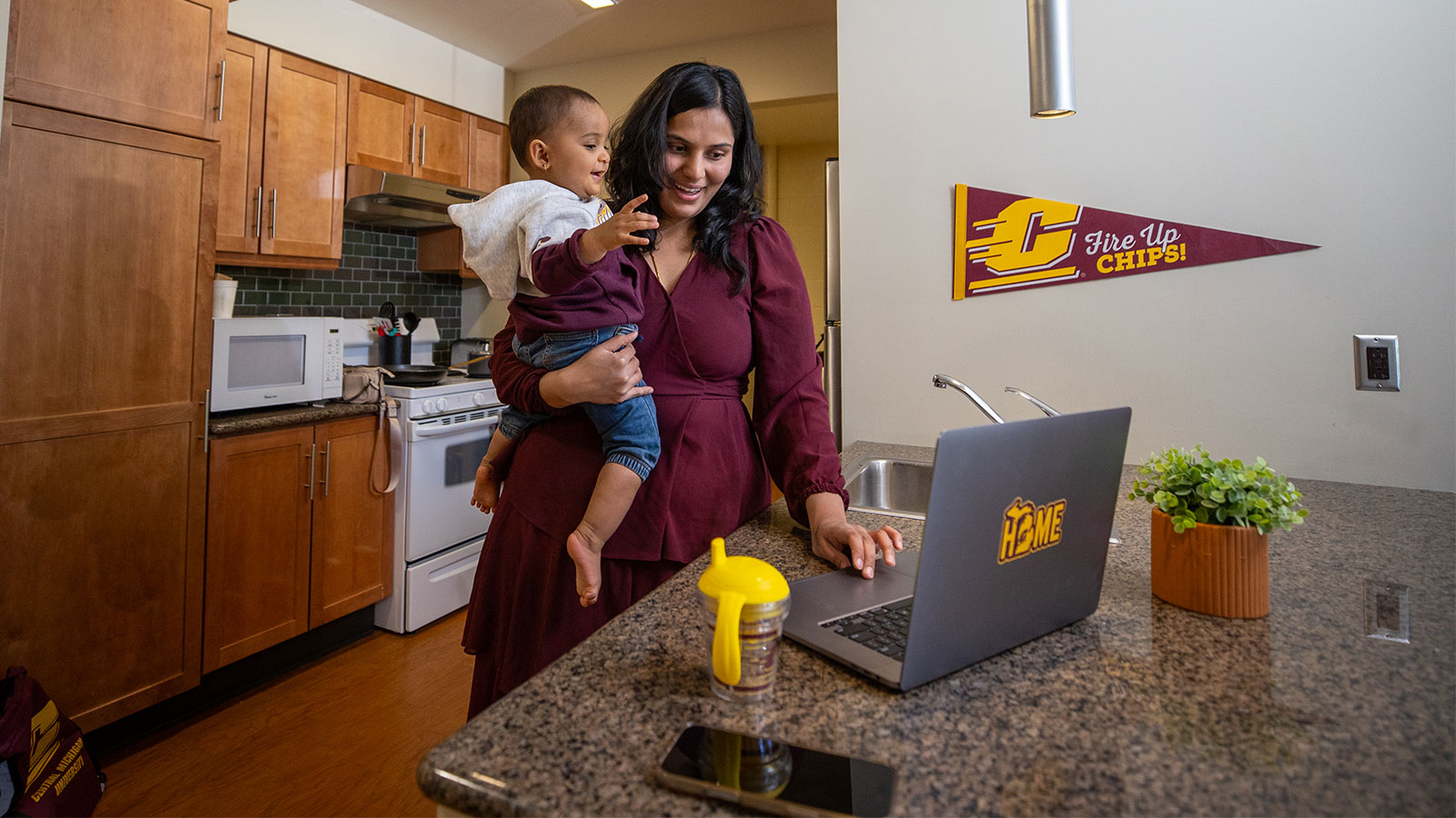 a woman in CMU's graduate housing, holding a baby in her arms while using a laptop with a 'fire up chips' flag on the wall