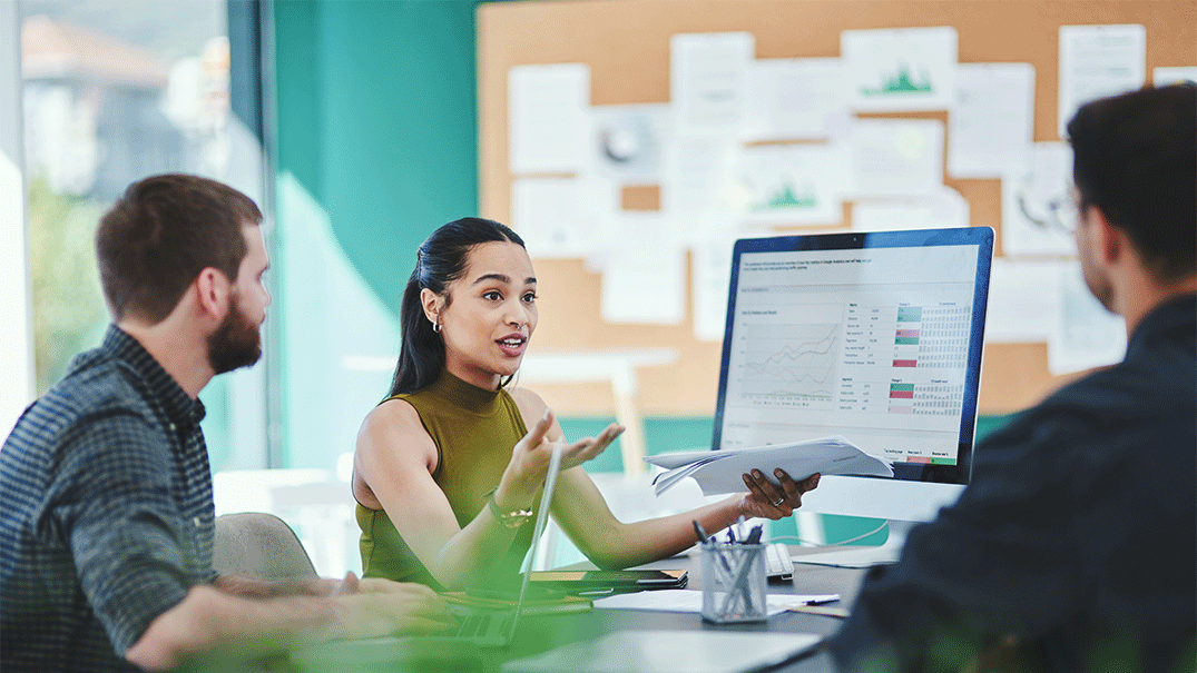 A woman with documents in one hand discussed statistics on a desktop screen with two men.