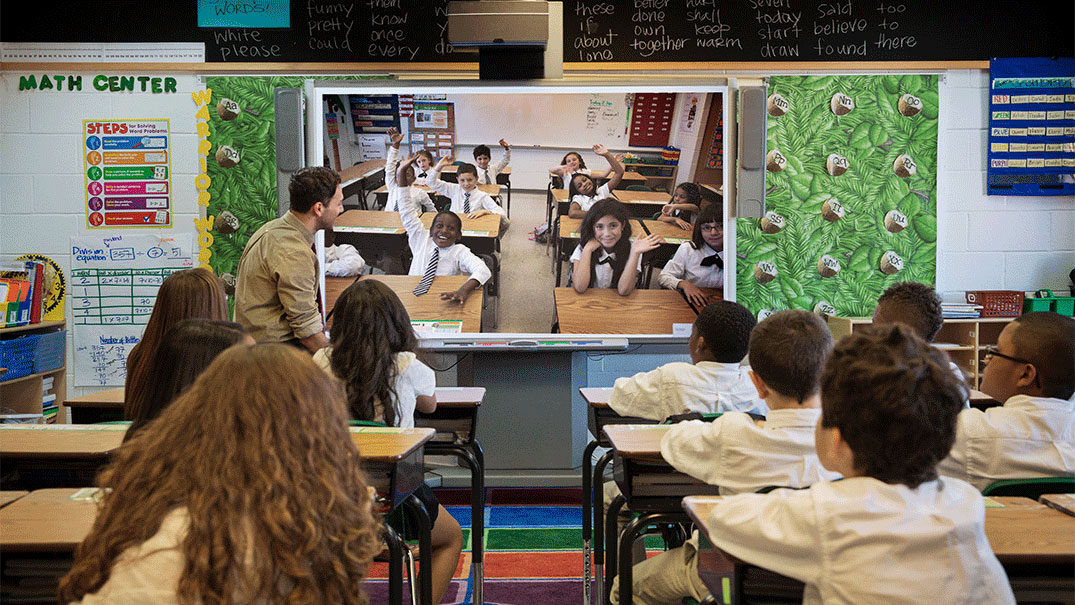 A teacher and a class full of students watching a monitor streaming another classroom with students waving