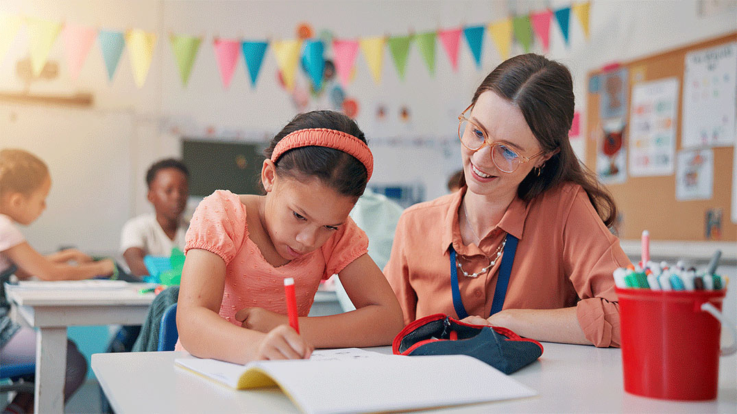 a teacher helps a young student write, with two children in the background