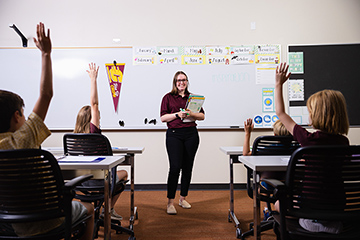 Teacher stands at the front of her classroom in CMU polo shirt as her students raise their hands to ask questions.