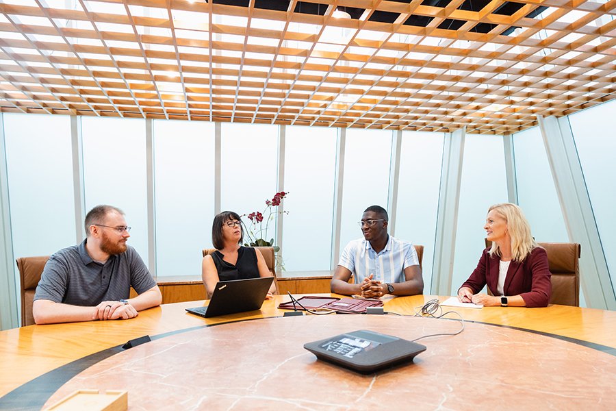 Four DET faculty members hold a meeting with online students. They are sitting in a large open conference area talking with unseen online students.