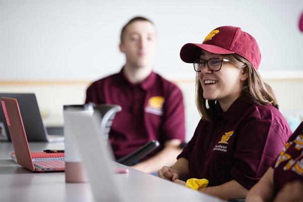A student wearing a baseball cap sits at a table with a laptop in front of her.