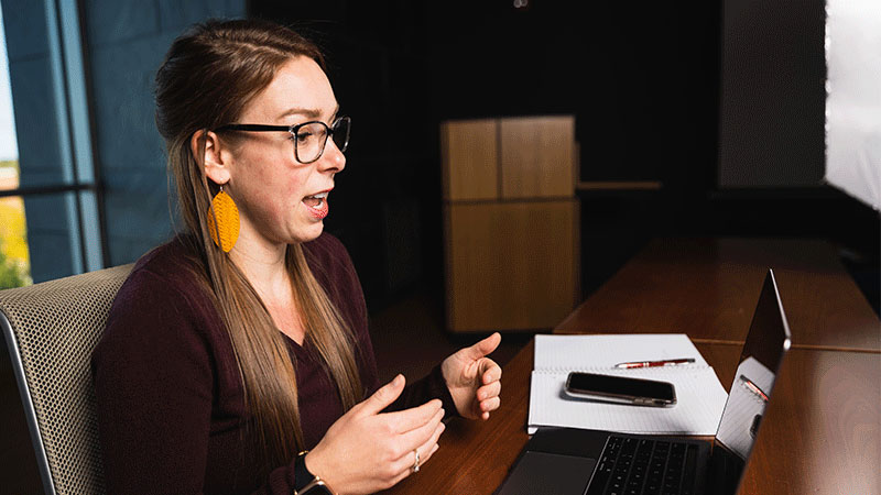 Woman speaking at her desk in front of her laptop next to a notebook and cellphone