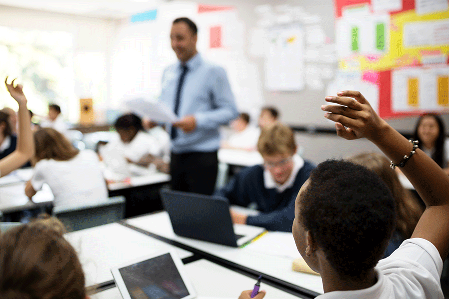 High school teacher walks among desks in his classroom. Students raise their hands to ask questions.